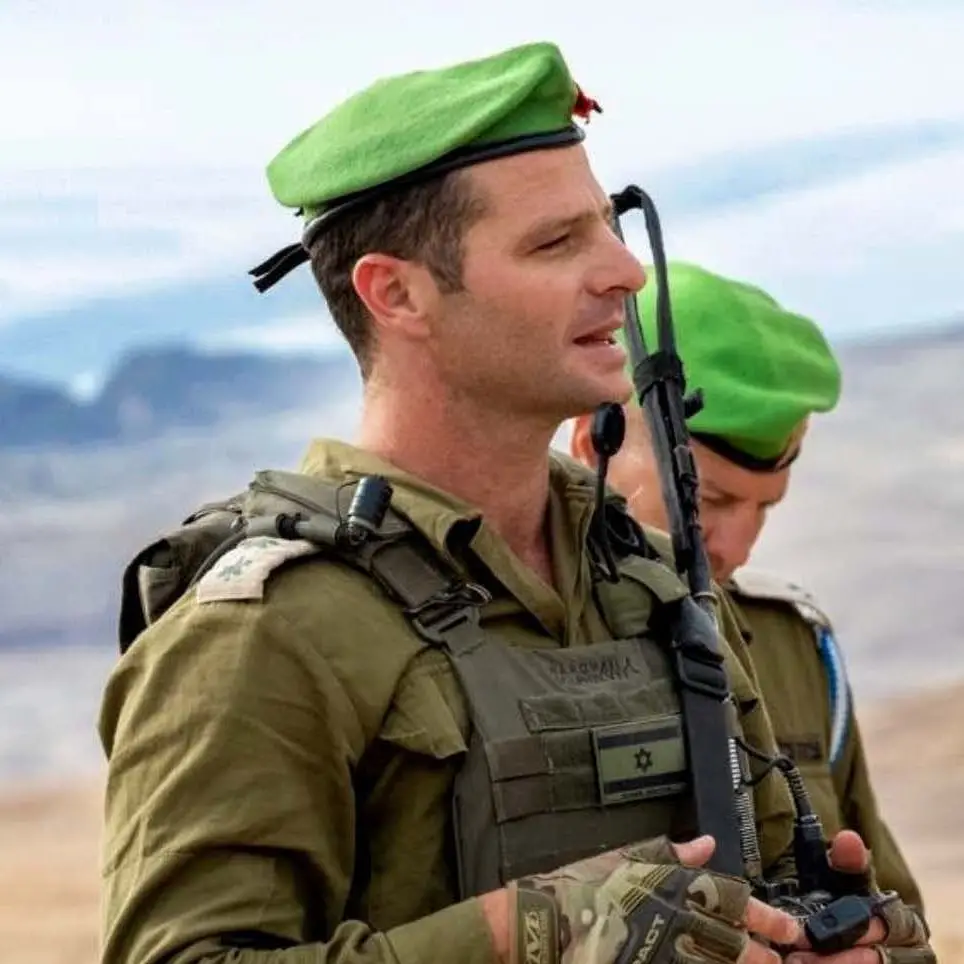 ברנש במדים ובכומתה ירוקה, עומד בשטח פתוח ומבטו לצד. מאחוריו לוחם נוסף, מטושטש.